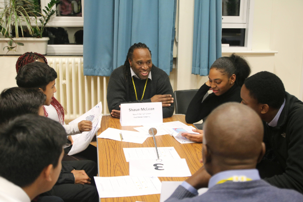 Six mentee's sit around a taable holding and looking at papers. in the centre sat at the head of the table is a Role Model smiling whilst talking to the students. In the foreground id the back of the head of another Role Model.