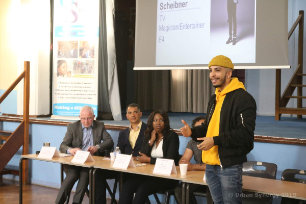 A school hall with a stage and screen in the background, in front of the stage is a row of tables with Role Models sat behind them, they are watching the person talking in front of the desk. The person talking is Troy the magician, he wears a yellow beanie and hoodie with a black jacket.