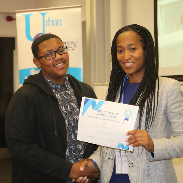 Leila shakes the hand of a mentee at their graduation. In Leila's left hand she holds a certificate, she wears a blue dress and grey blaxer. The mentee is wearing a black and white top with an abstract pattern, and a black zip up jumper over the top.
