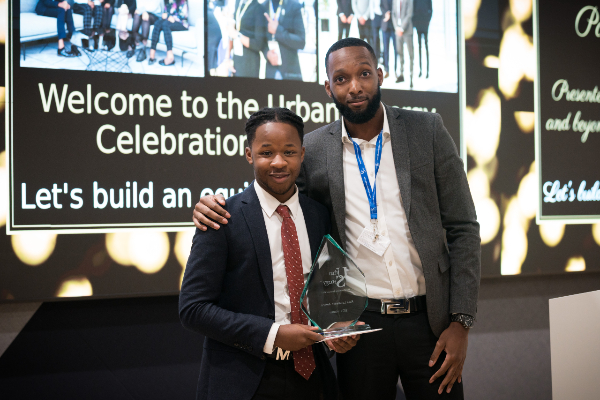 In the centre of the frame stand Nathaneal receiving the first ever Paul Lawrence Award at the 2021 Urban Synergy Celebration Event. Presenting the award is Kaream Lawrence, Paul's son. Nathaneal wears a blue suit with a red and white polka dot tie. Kaream wears a white shirt with a grey suit jacket and a blue lantard around his neck. In the background there is a screen with a presentation.