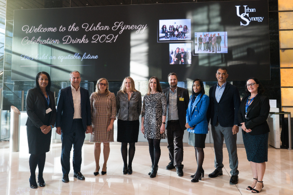 nine people stand in the entrance of a large room with marble floors. In the background is a screen for the Urban Synergy Celebration event 2021. Nine individuals stand in a line in front of the screen, including Urban Synergy CEO and Founder Leila Thomas, other Urban Synergy staff and corporate sponsors.